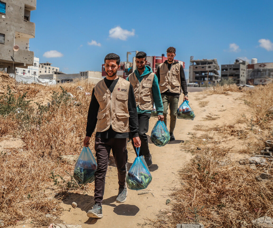 photo of three men walking down a dirt hill holding blue plastic bags of aid. 