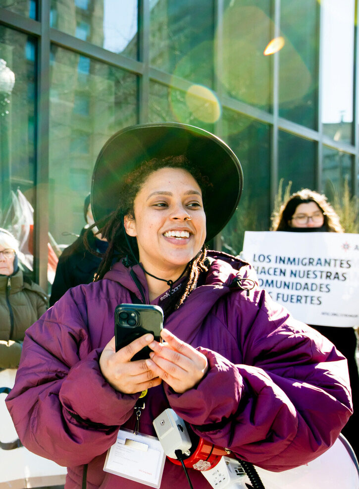 Color photo of woman in big puffy purple jacket, holding her phone and a megaphone smiling to the side wearing a wide brim hat. In the back someone holds a sign that says 
