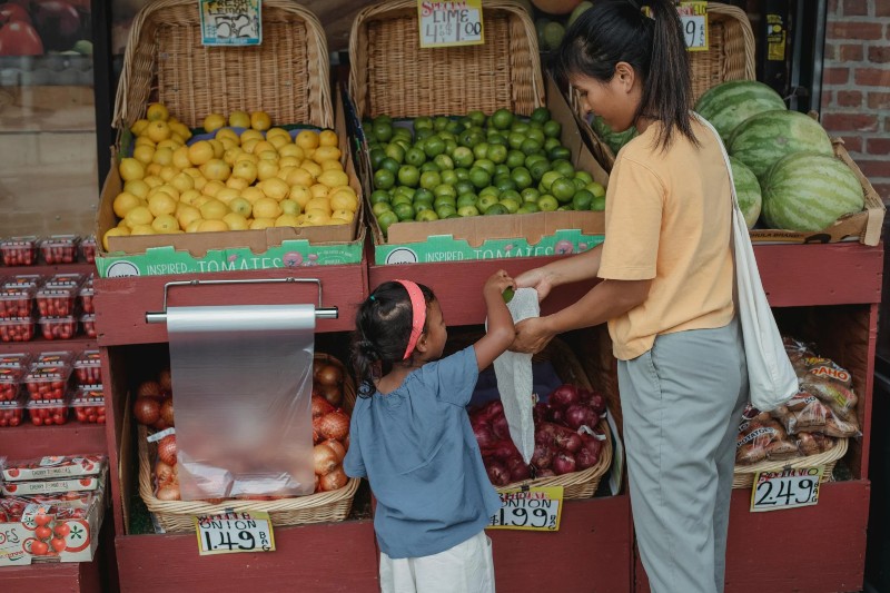 A woman and girl stand in front of a produce stand. The woman holds a plastic bag as the girl puts produce into it.