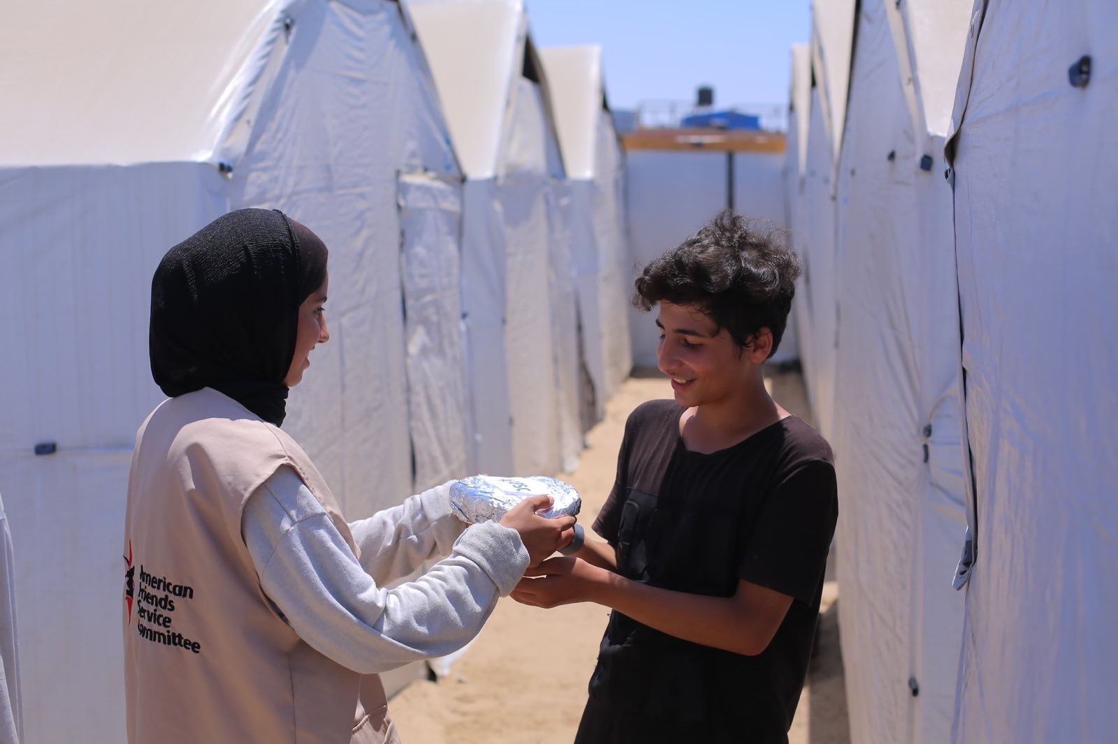Woman in AFSC vest hands a package of food to a boy among a row of tents