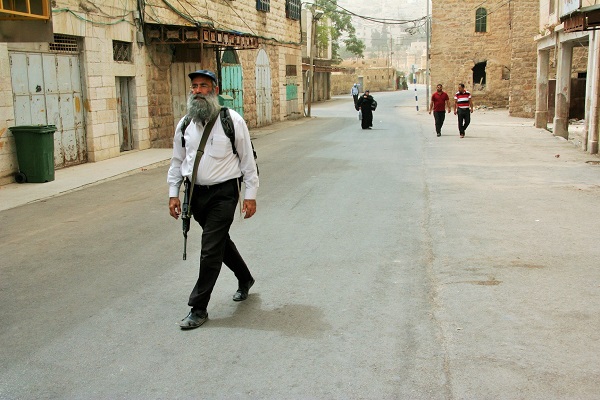 An armed Israel settler who was chaperoning a group of teenage youth walks towards the gathering site where dozens of settlers are disrupting the call to prayer.